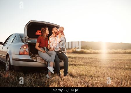 Joyeux jeune maman et papa de famille avec leur petit fils en appréciant pique-nique de week-end d'été assis sur le tronc de la voiture à l'extérieur de la ville dans le champ à Banque D'Images