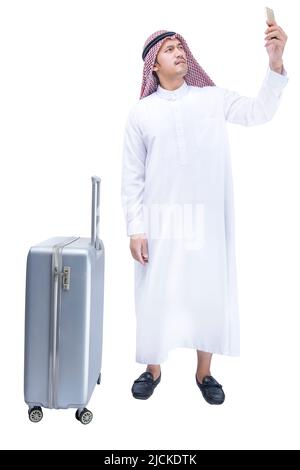 Homme arabe portant le keffiyeh regardant un téléphone portable avec une valise pour voyager isolé sur fond blanc Banque D'Images