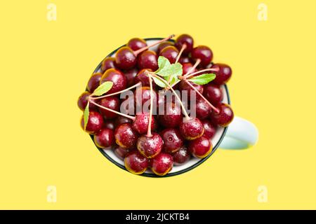 Gros plan d'une tasse rustique remplie de cerise mûre rouge fraîche avec des branches et des gouttes d'eau au centre, vue du dessus sur fond jaune. Fruits doux. Banque D'Images