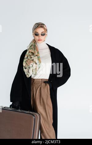 femme en manteau noir, lunettes de soleil tendance et mouchoir debout avec valise vintage isolée sur gris Banque D'Images