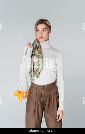 femme en col roulé blanc et foulard portant un sac à filet avec des oranges mûres isolées sur le gris Banque D'Images