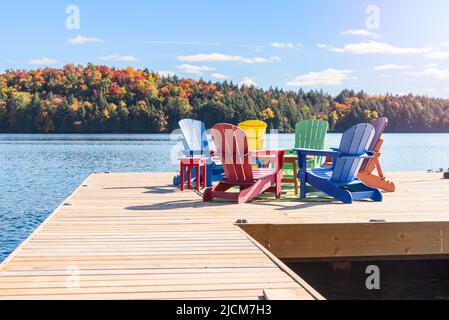 Chaises adirondack colorées autour d'une table sur une jetée en bois sur un lac par un jour d'automne clair. Banque D'Images