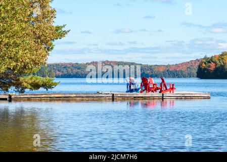 Chaises adirondack colorées sur une jetée flottante sur un lac avec des rives boisées par un beau jour d'automne. Feuillage d'automne. Banque D'Images