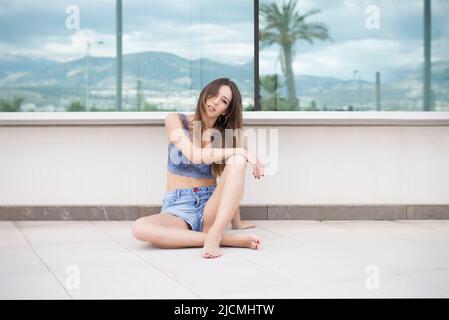Vue latérale complète de la jeune femme dans des vêtements décontractés assis au sol près de la fenêtre et regardant l'appareil photo Banque D'Images