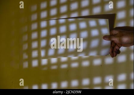 Homme mature tient un morceau de papier blanc rectangulaire dans sa main. Répétition d'un motif géométrique de carrés de lumière sur un mur jaune. Lumière abstraite a Banque D'Images