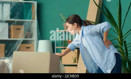 Une femme ressent une douleur soudaine au dos lorsqu'elle tente de soulever une boîte à carton. Banque D'Images