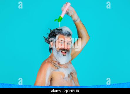 Homme barbu souriant versant du shampooing sur la tête. Soins capillaires pour hommes. Lavage des cheveux. Prendre une douche. Heure du bain. Banque D'Images