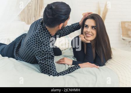 Homme adulte aux cheveux sombres et belle femme smiley allongé sur leur lit en vêtements. Le mari touche les cheveux longs et lisses de sa femme. Photo de haute qualité Banque D'Images
