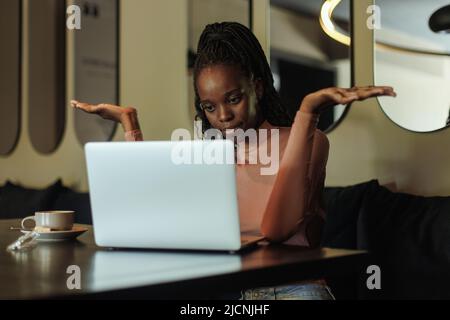 Une femme multiraciale perplexe qui boit du café, travaille avec un ordinateur portable, dans un café, se lance dans la main. Problème de tâche à distance, je ne sais pas Banque D'Images