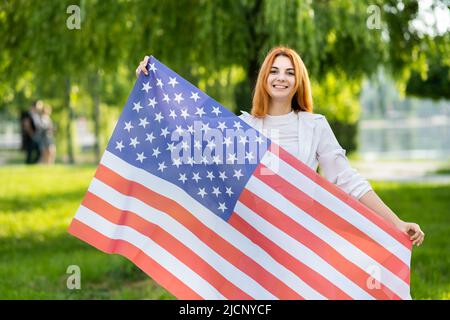Bonne jeune femme aux cheveux rouges posant avec le drapeau national des États-Unis debout à l'extérieur dans le parc d'été. Fille positive avec bannière États-Unis dehors Banque D'Images
