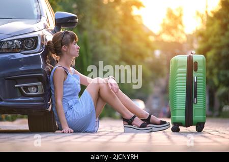 Jeune femme fatiguée avec une valise assise près de sa voiture attendant quelqu'un. Concept de voyage et de vacances Banque D'Images
