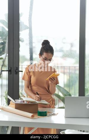 Belle femme entrepreneur préparant des boîtes de colis et des marchandises d'emballage tout en vérifiant les commandes sur son mobile Banque D'Images