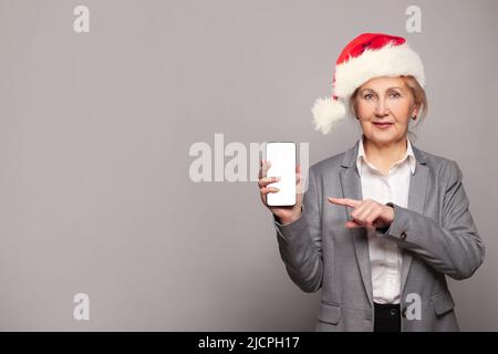 Bonne femme mûre souriante en chapeau de père Noël rouge tenant un smartphone avec un écran vide Banque D'Images