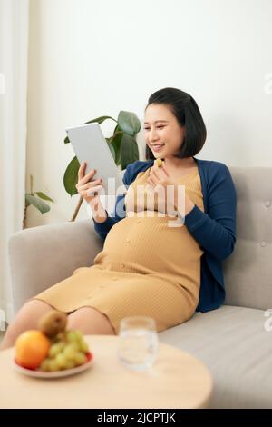 Bonne jeune femme enceinte asiatique mangeant des fruits et tenant un comprimé tout en étant assise sur un canapé à la maison. Banque D'Images