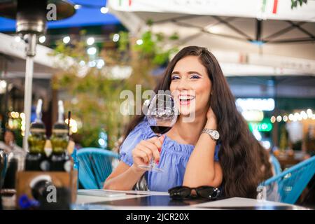 Une jeune femme au maquillage lumineux boit du vin d'un verre de vin dans un café de rue et rit, regardant la caméra. Banque D'Images