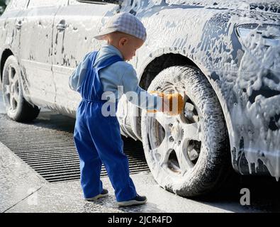 Petit garçon debout près de l'auto recouvert de mousse, tenant l'éponge dans sa main et apprenant à laver la voiture. Enfant de sexe masculin étudiant pour laver l'automobile avec une éponge, se pliant vers la roue avant. Banque D'Images