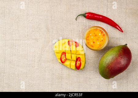 Vue de dessus de la mangue entière, du piment entier, du piment en tranches, de la moitié coupée de la mangue et du chutney à la mangue épicée dans un bol en verre sur le jute avec place pour te Banque D'Images