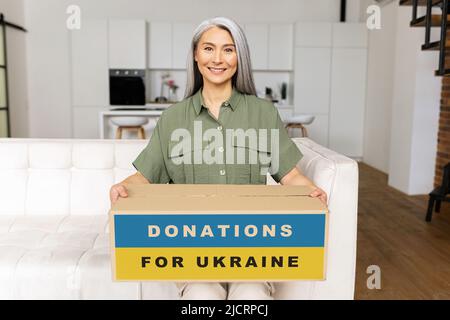 Une femme asiatique de haut niveau souriante, assise sur un canapé, emballant des marchandises dans une boîte en carton pour les Ukrainiens, faisant un don à une œuvre caritative, les gens du monde entier soutiennent les réfugiés d'Ukraine Banque D'Images