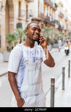Ravi jeune Afro-américain élégant homme dans t-shirt et dans l'ensemble sourire. Il regarde loin tout en parlant sur un smartphone, debout dans la rue de la ville avec la main dans la poche Banque D'Images