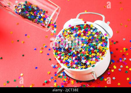 Un verre aux sequins multicolores, un réveil et une cravate en forme d'étoile sur fond rouge. Fête, nouvel an, célébration concept Banque D'Images