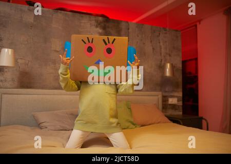 Portrait d'un enfant insouciant méconnaissable jouant avec une boîte en carton et sautant sur le lit à la maison Banque D'Images