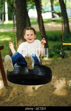 Bonne petite fille balançant sur la roue en caoutchouc de voiture dans l'aire de jeux dans le parc vert. Vacances d'été dans le camp, centre touristique. Marcher et jouer à l'extérieur Banque D'Images