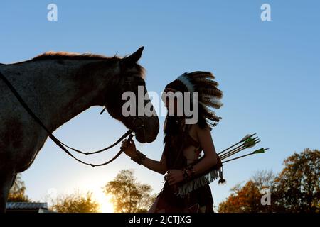 Une jeune fille est vue au coucher du soleil comme un Indien amérindien. Elle est vue avec son cheval comme le soleil se met dans la distance. Banque D'Images