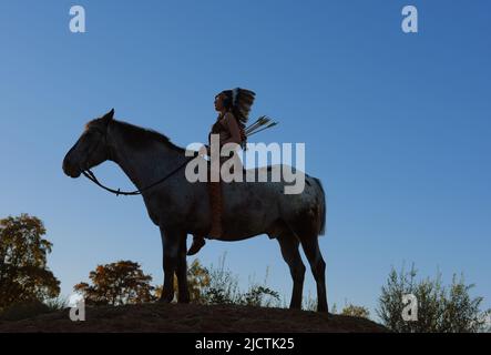 Une jeune fille est vue au coucher du soleil comme un Indien amérindien. Elle est vue avec son cheval comme le soleil se met dans la distance. Banque D'Images