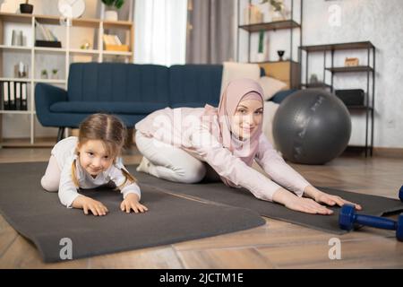 Belle mère arabe dans hijab et charmante petite fille sont souriants tout en pratiquant le sport ensemble à la maison, faisant de l'exercice de pose d'enfant. Banque D'Images