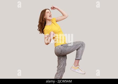 Pleine longueur excitée jeune fille en décontracté jaune T-shirt et jeans gris posant, accrochant poings et hurler heureux, célébrant son triomphe. Prise de vue en studio isolée sur fond gris. Banque D'Images