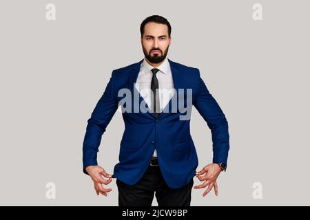 Portrait d'un triste barbu malheureux montrant des poches vides à l'intérieur, allant en faillite, pas d'argent pour la vie, portant un costume de style officiel. Prise de vue en studio isolée sur fond gris. Banque D'Images