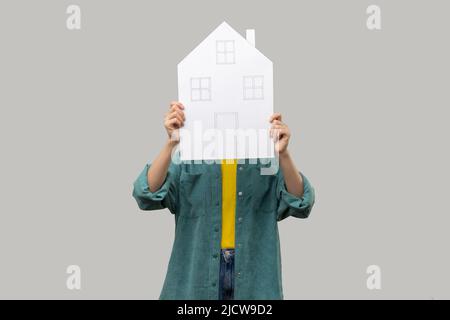 Une femme inconnue se cache derrière une maison en papier avec des fenêtres et des portes dessinées, un programme de logement gouvernemental, rêver d'une nouvelle maison, porter une veste de style décontracté. Prise de vue en studio isolée sur fond gris Banque D'Images