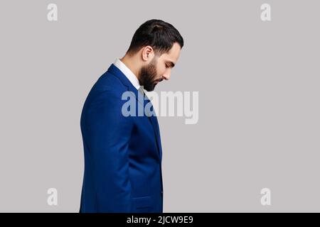 Portrait d'un triste homme barbu debout avec sa tête vers le bas et regardant, ayant avec une tristesse insatisfaite visage, portant un costume de style officiel. Prise de vue en studio isolée sur fond gris. Banque D'Images