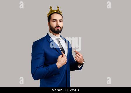 Portrait d'un homme barbu plein de confiance en lui-même, en couronne dorée, pointant vers elle-même avec le doigt, statut privilégié supérieur, portant un costume de style officiel. Prise de vue en studio isolée sur fond gris. Banque D'Images