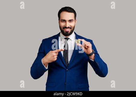 Portrait d'un homme d'affaires barbu ravi et satisfait tenant le doigt pointant sur le bitcoin d'or, ecommerce, portant un costume de style officiel. Prise de vue en studio isolée sur fond gris. Banque D'Images