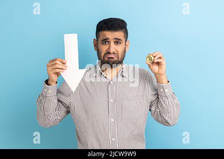 Portrait d'un homme d'affaires malheureux montrant du bitcoin et une flèche blanche pointant vers le bas, exprimant des émotions négatives, portant une chemise rayée. Studio d'intérieur isolé sur fond bleu. Banque D'Images