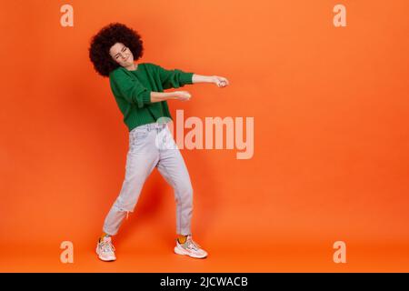 Portrait de profil complet de la femme avec la coiffure afro dans le chandail vert tirant invisible lourd fardeau, s'efforçant dur pour atteindre le succès. Studio d'intérieur isolé sur fond orange. Banque D'Images