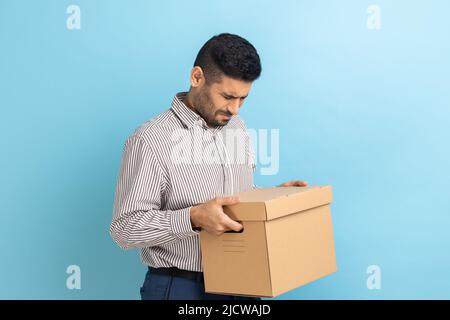 Portrait d'un travailleur triste tenant une parcelle de carton, contrarié d'être tiré au travail, pleurant, exprimant la tristesse, portant une chemise rayée. Studio d'intérieur isolé sur fond bleu. Banque D'Images