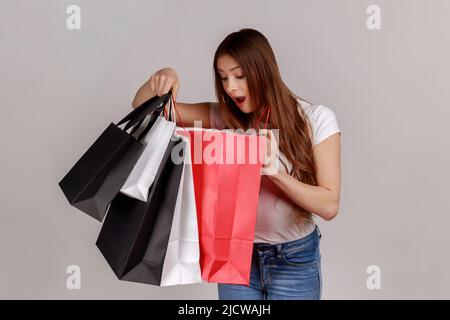 Portrait d'une femme excitée tenant regardant à l'intérieur des sacs de shopping, étant étonné avec l'achat bon marché ou d'un excellent cadeau, portant T-shirt blanc. Prise de vue en studio isolée sur fond gris. Banque D'Images