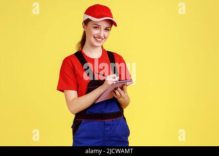 Portrait d'une femme souriante et heureuse qui écrit dans un carnet papier, qui fait une liste de tâches, écrit des commandes, porte une combinaison et une casquette rouge. Studio d'intérieur isolé sur fond jaune. Banque D'Images