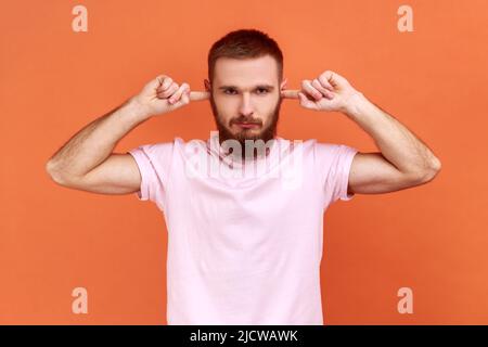 Portrait de l'homme barbu ferme les oreilles avec les doigts regardant sérieusement l'appareil photo, fatigué du bruit irrité, veut le silence, portant un T-shirt rose. Studio d'intérieur isolé sur fond orange. Banque D'Images