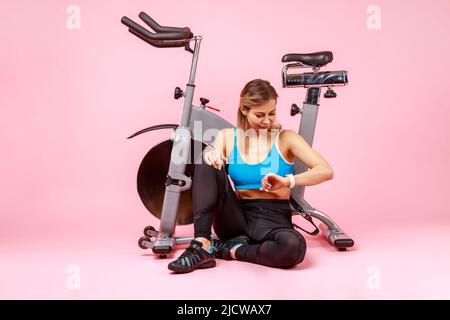 Portrait complet d'une femme athlétique regardant son tracker d'activité physique, vérifiant les indicateurs après l'entraînement, portant des collants de sport et un haut. Studio d'intérieur isolé sur fond rose. Banque D'Images