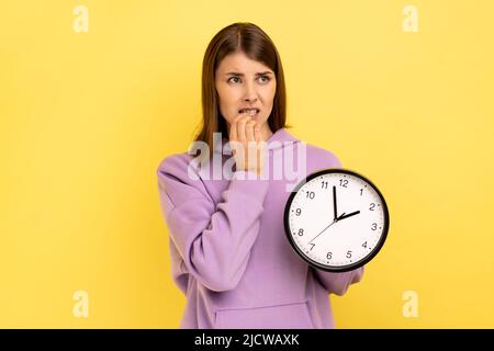 Portrait de la femme nerveuse impatiente attirante mordant ses ongles et tenant une grande horloge murale, échéance, regardant loin, portant le sweat à capuche violet. Studio d'intérieur isolé sur fond jaune. Banque D'Images