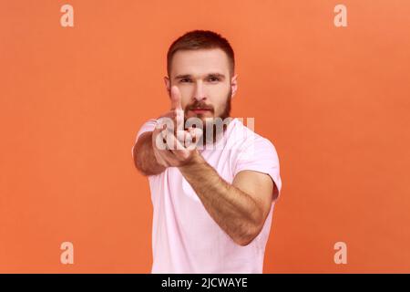 Portrait d'un homme barbu courageux tenant les doigts prétendant tenir le fusil, visant l'ennemi, auto-défense, portant un T-shirt rose. Studio d'intérieur isolé sur fond orange. Banque D'Images