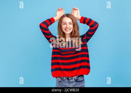 Portrait d'une femme gaie portant un chandail rayé de style décontracté montrant le geste des oreilles de lapin et regardant ludique heureux, comportement puéril. Studio d'intérieur isolé sur fond bleu. Banque D'Images