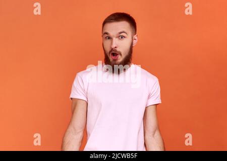 Portrait de l'homme barbu stupéfié debout avec la bouche ouverte dans la surprise, a choqué l'expression, entend des nouvelles incroyables, portant le T-shirt rose. Studio d'intérieur isolé sur fond orange. Banque D'Images