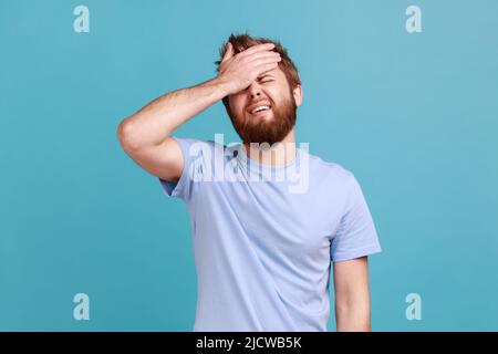 Portrait de bouleversé triste barbu homme debout avec geste de facipalm, se blâmant, sentiment de tristesse regret à cause de la mauvaise mémoire. Studio d'intérieur isolé sur fond bleu. Banque D'Images