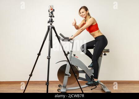 Femme athlétique enregistrant vidéo tutoriel sur téléphone portable sur trépied, vélo d'entraînement et montrant le pouce vers le haut, portant des collants de sport et haut rouge. Studio d'intérieur tourné sur fond de mur gris. Banque D'Images