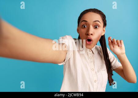 Femme choquée avec des dreadlocks faisant le selfie, regardant l'appareil photo avec les grands yeux et la bouche ouverte, point de vue photo, portant une chemise blanche. Studio d'intérieur isolé sur fond bleu. Banque D'Images