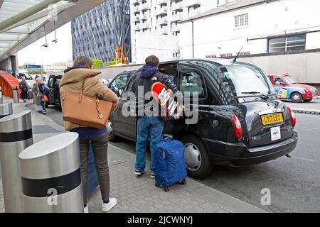 Jeune couple vue arrière sortir d'un taxi noir avec bagages et planche à roulettes à la gare de Paddington à Londres Angleterre Royaume-Uni 2022 Banque D'Images